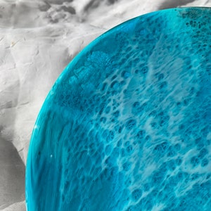 May include: A close-up of a round, ocean-themed resin art piece. The artwork features shades of turquoise and white, mimicking the look of ocean waves. The resin has a glossy finish, with a textured, bubbly pattern.