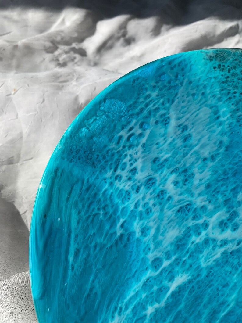 May include: A close-up of a round, ocean-themed resin art piece. The artwork features shades of turquoise and white, mimicking the look of ocean waves. The resin has a glossy finish, with a textured, bubbly pattern.