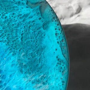 May include: Close-up of a round, resin art piece with a swirling ocean-like design. The artwork features shades of turquoise, teal, and white, creating a textured, wave-like effect. The edges are smooth and clear, showcasing the depth of the colors.