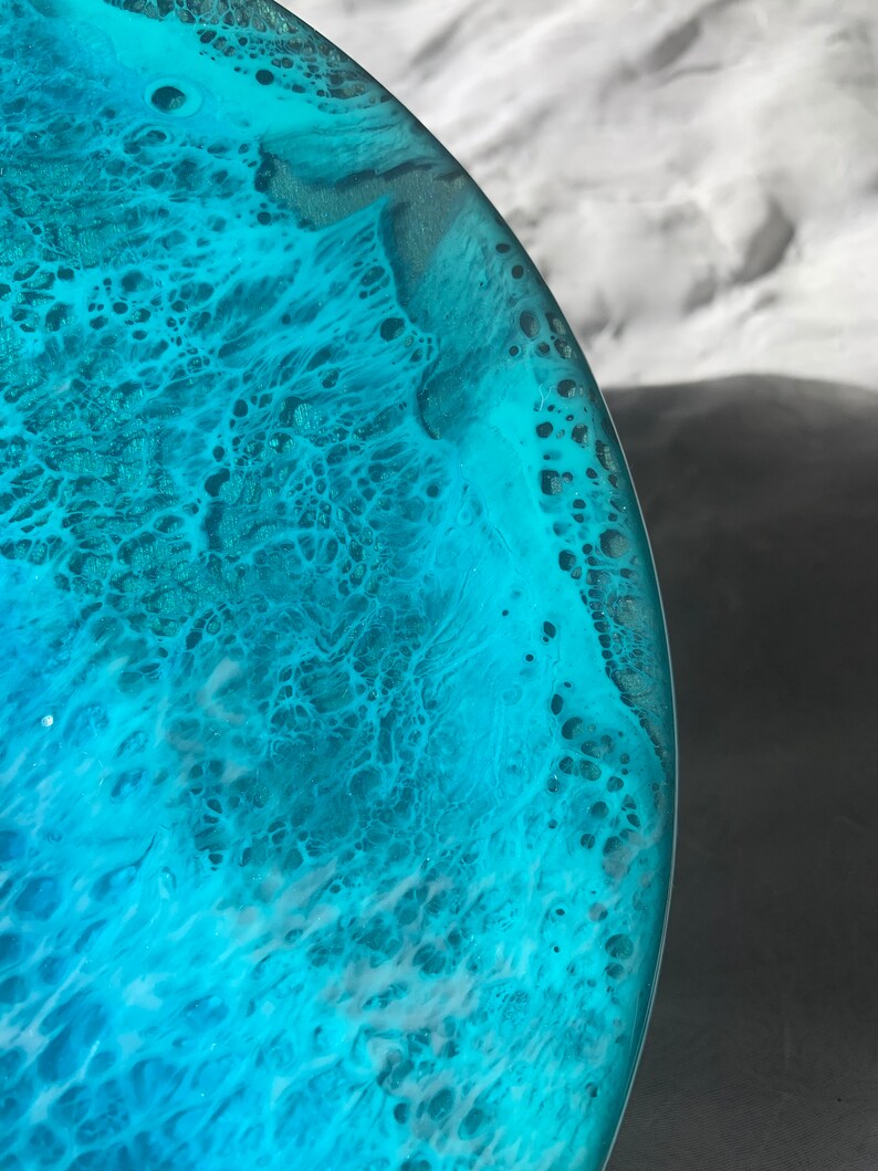 May include: Close-up of a round, resin art piece with a swirling ocean-like design. The artwork features shades of turquoise, teal, and white, creating a textured, wave-like effect. The edges are smooth and clear, showcasing the depth of the colors.