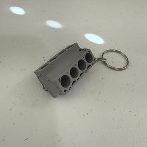 May include: A gray, miniature engine block keychain. The keychain features a detailed replica of an engine block with multiple cylinders and a key ring attached. The item is made of a solid, gray material and is designed to be a functional and decorative accessory.