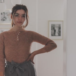 May include: A woman wearing a light pink crocheted sweater with pearl buttons and a gray skirt. She is standing in a room with white walls and a picture on the wall behind her.