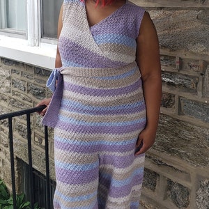 May include: A woman wearing a long, striped crochet dress with a V-neck and a tie at the waist. The dress is in shades of purple, blue, and beige.
