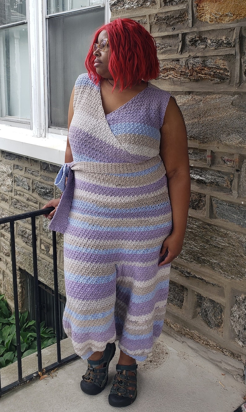 May include: A woman wearing a long, striped crochet dress with a V-neck and a tie at the waist. The dress is in shades of purple, blue, and beige.