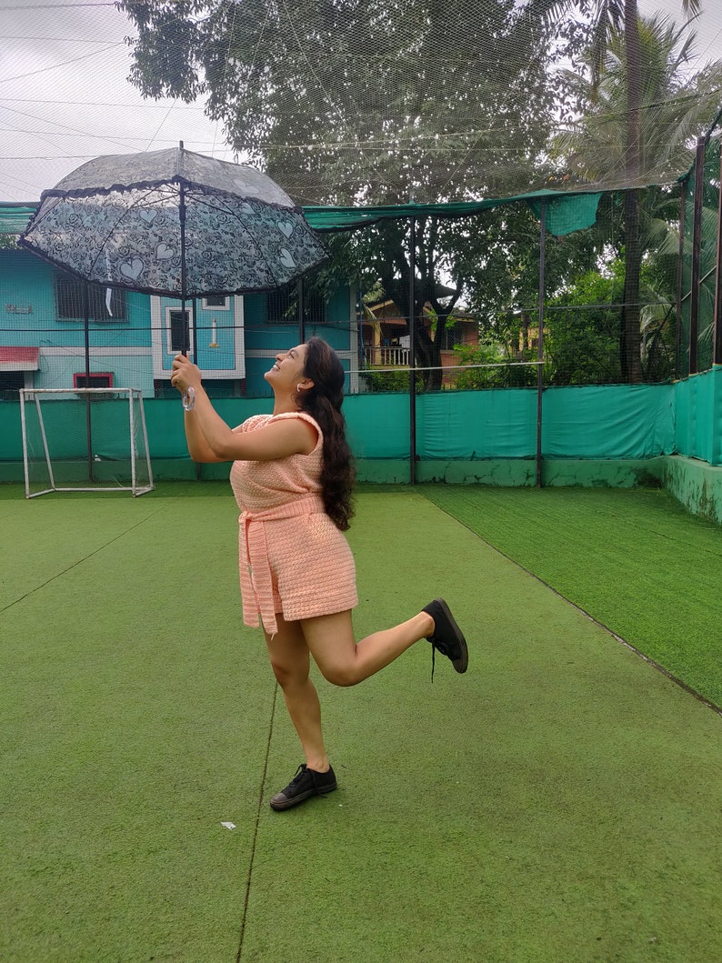 May include: A woman in a pink dress holds a black and white lace umbrella while standing on a green artificial grass field.