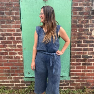 May include: A woman wearing a blue crocheted jumpsuit with a wide belt. She is standing in front of a green wall.