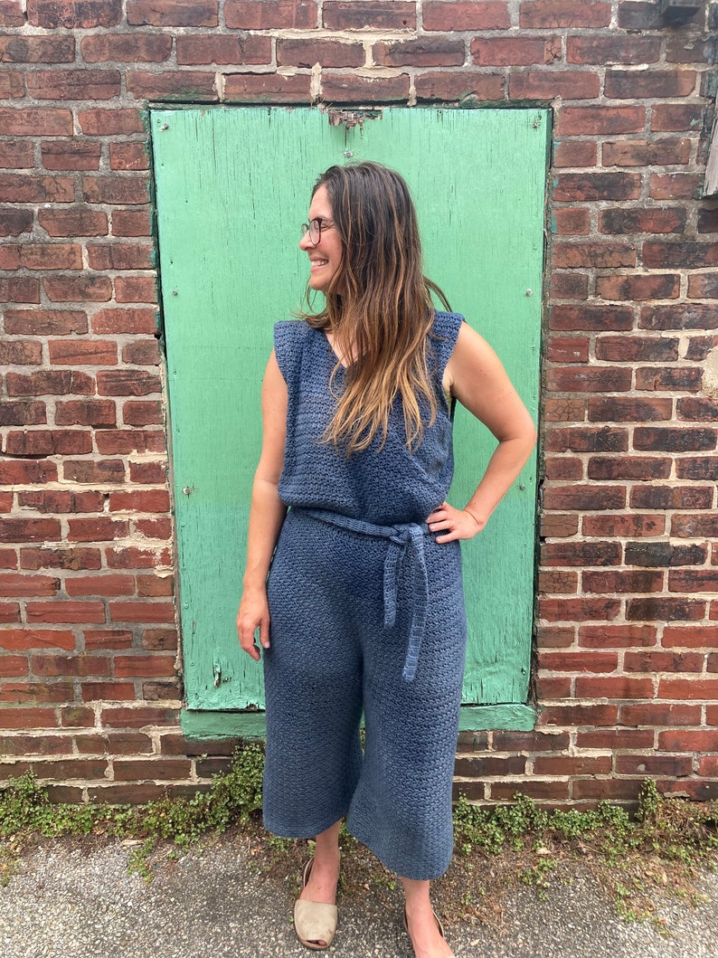 May include: A woman wearing a blue crocheted jumpsuit with a wide belt. She is standing in front of a green wall.
