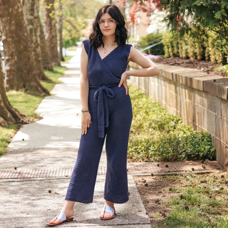 May include: A woman wearing a navy blue crochet jumpsuit with a wide belt. She is standing on a sidewalk with trees in the background.