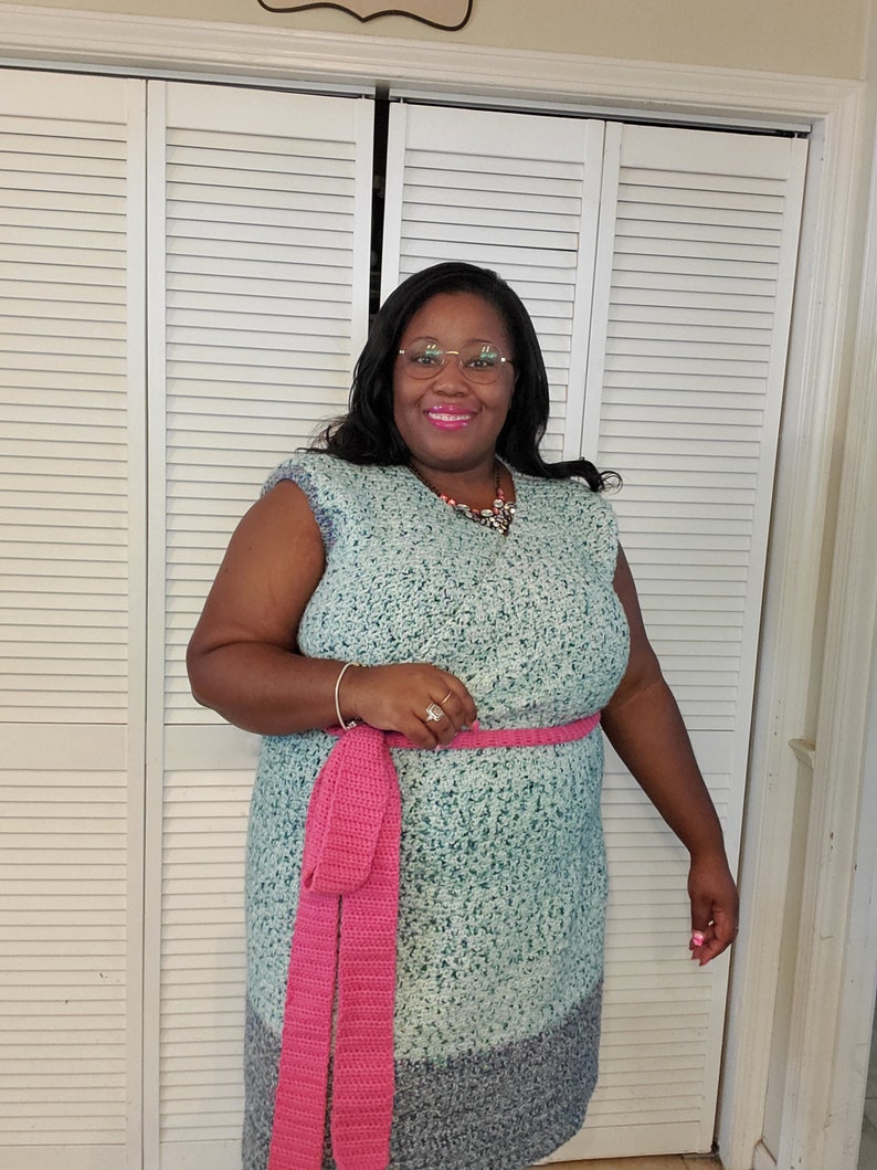 May include: A teal blue crochet dress with a pink crocheted belt. The dress has a sleeveless design and a slightly flared skirt.