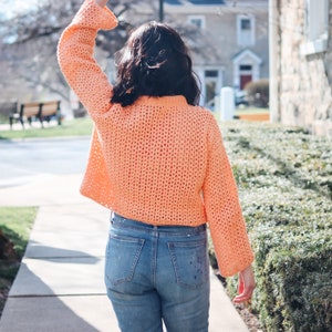 Könnte beinhalten: Eine Frau trägt einen peachfarbenen, locker sitzenden Strickpullover mit langen Ärmeln. Sie trägt eine blaue Jeans mit weißen Farbspritzern.