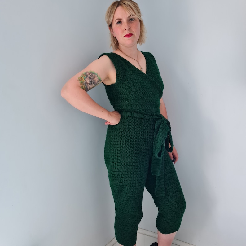 May include: A dark green crochet jumpsuit with a V-neck and a self-tie belt. The jumpsuit has a textured, crocheted fabric.