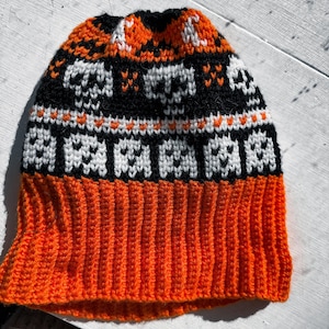 Könnte beinhalten: Eine schwarze, weiße und orangefarbene Strickmütze mit Totenkopf- und Geistermuster. Die Mütze hat einen gerippten Bund.