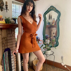 May include: A woman wearing a rust-colored crochet mini dress with a tie belt. The dress has a V-neckline and sleeveless design. She is standing in a room with a fireplace and a mirror.