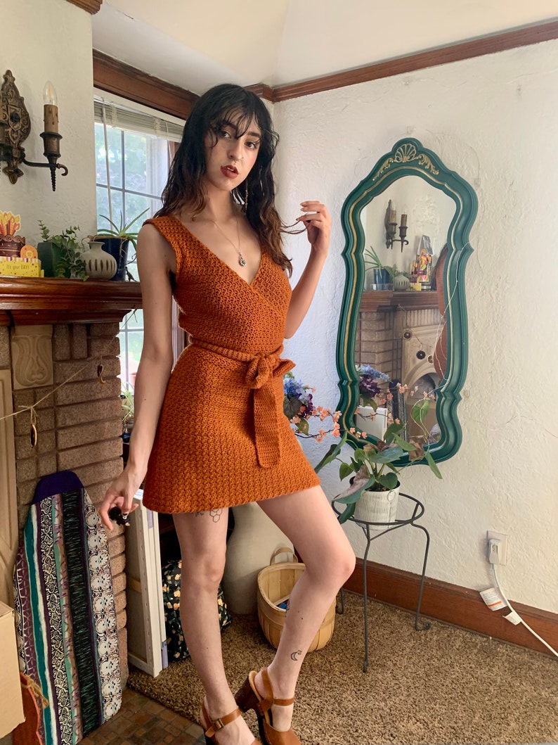 May include: A woman wearing a rust-colored crochet mini dress with a tie belt. The dress has a V-neckline and sleeveless design. She is standing in a room with a fireplace and a mirror.