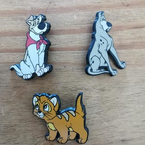 May include: Three cartoon character pins featuring a white dog with a red bandana, a grey dog with black spots, and an orange tabby cat.
