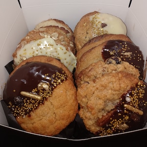 May include: A box of assorted cookies with chocolate and white icing and gold sprinkles. The cookies are in a white box with a black interior.