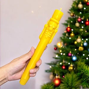 Puede incluir: Una vela amarilla con forma de cascanueces. La vela es alta y delgada, con un sombrero puntiagudo y una cara detallada. La vela se sostiene en una mano, con un árbol de Navidad en el fondo.