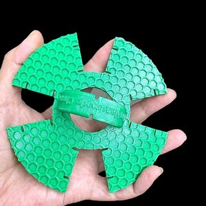 Könnte beinhalten: Ein hellgrünes, 3D-gedrucktes Objekt mit Wabenmuster. Das Objekt wird in einer Hand vor schwarzem Hintergrund gehalten. Es ist kreisförmig mit vier fächerartigen Abschnitten und einem zentralen Ring mit dem Text "Handcrafted Club".