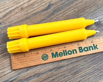 May include: Two yellow taper candles with a ribbed base, measured against a wooden ruler with the text "Mellon Bank" printed on it.