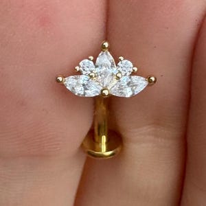 May include: A gold-colored lip piercing stud with a floral design. The stud features a cluster of clear, marquise-cut gemstones arranged in a flower-like pattern. The post is a simple, straight design.