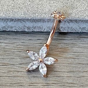 May include: Rose gold belly button ring with a clear flower design. The flower has five petals with clear stones.