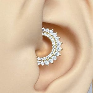 Könnte beinhalten: Ein goldfarbener, kreisförmiger Ohrring mit einem Kranz aus klaren, funkelnden Steinen. Der Ohrring wird in einem menschlichen Ohrmodell präsentiert, um sein Design hervorzuheben. Das Schmuckstück ist für das Ohr gedacht.