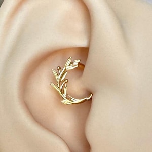 May include: A gold-colored ear piercing hoop with a delicate leaf design. The jewelry is set in a human ear, showcasing the intricate details of the floral-inspired adornment. The hoop is designed to be worn in the ear's cartilage.