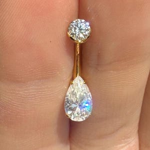 May include: A gold-coloured belly button ring with two clear, sparkling gemstones. The top stone is round, and the bottom stone is a pear shape. The jewellery is held up by a thin, gold-coloured bar.