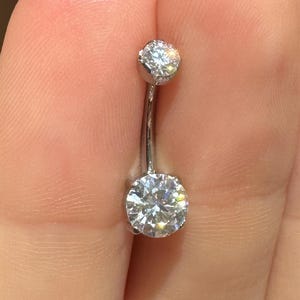 May include: A silver belly button ring with two clear, round-cut gemstones. The larger gemstone is at the bottom, and the smaller one is at the top. The curved bar is visible between the gems.