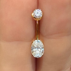 May include: A gold-colored belly button ring featuring two clear, sparkling gemstones. The top stone is round, while the lower stone is an oval shape. The ring is held between fingers, showcasing its delicate design.