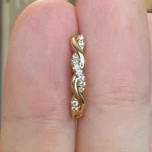Puede incluir: Un anillo dorado con un diseño retorcido, adornado con pequeñas piedras preciosas claras. El anillo se sostiene entre dos dedos, mostrando sus intrincados detalles y brillo. El diseño del anillo presenta un patrón trenzado, con las piedras preciosas engarzadas a lo largo de los giros.
