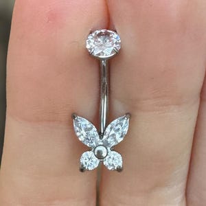 May include: A silver belly button ring with a butterfly design. The ring features a large, round, clear gem at the top and a butterfly made of smaller, clear gems at the bottom. The butterfly has two marquise-shaped gems for wings and two round gems for the body.