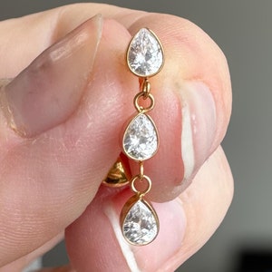Könnte beinhalten: Ein goldfarbener Ohrring mit drei klaren, tropfenförmigen Edelsteinen. Die Edelsteine sind vertikal angeordnet und erzeugen einen Kaskadeneffekt. Der Ohrring wird von einer Hand gehalten, wodurch das Design und der Glanz des Schmucks hervorgehoben werden.