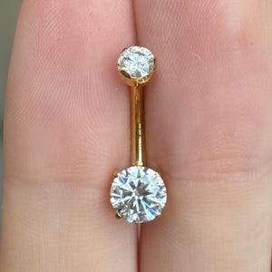 May include: A gold-colored belly button ring with two clear, round, sparkling gemstones. The top gemstone is smaller than the bottom one. The jewelry is held between fingers against a blurred background.