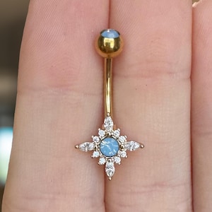 May include: A gold-colored belly button ring with a starburst design. The ring features a light blue center stone surrounded by clear, sparkling accents. The top ball also has a light blue stone.