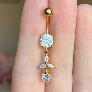 Puede incluir: Anillo de ombligo dorado con una gema redonda transparente y un dije colgante con tres gemas transparentes más pequeñas.