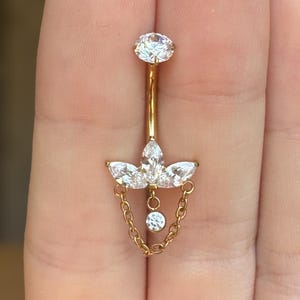 May include: A gold-colored belly button ring featuring a large, round, clear gemstone at the top. The ring has a decorative element with three marquise-cut gemstones and a dangling chain with a small, round gemstone at the end.