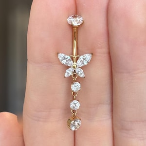 May include: A gold-coloured belly button ring with a butterfly design and clear, sparkling gemstones. The butterfly is the focal point, with a dangling chain of three additional gemstones. The top and bottom stones are round, while the butterfly's wings are marquise-shaped.