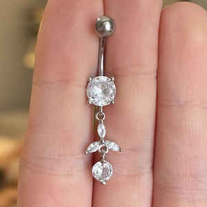 Puede incluir: Un piercing de ombligo plateado con un colgante de cristal transparente. El colgante tiene un cristal redondo con cristales más pequeños a cada lado.