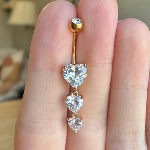May include: A gold-colored belly button ring featuring three clear, heart-shaped cubic zirconia stones of descending size. The top of the ring has a small, round clear stone. The jewelry is held in a hand.