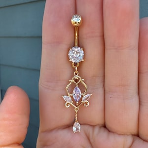 Puede incluir: Anillo de ombligo dorado con un dije colgante con un diseño floral y diamantes de imitación transparentes y morados.