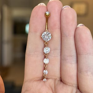 May include: A gold-coloured belly button ring with a curved barbell design. The ring features a series of clear, round-cut gemstones of varying sizes, suspended from a central gem. The top and bottom of the barbell have small, round gems.