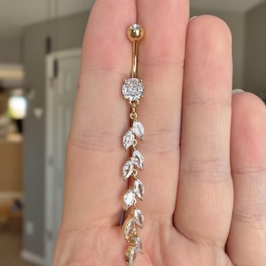 Puede incluir: Anillo de ombligo dorado con una cadena colgante de piedras de circonita cúbica transparentes en forma de hojas.