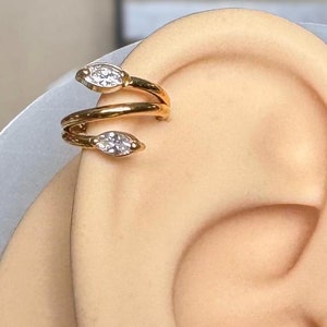 Puede incluir: Pendiente de oreja de oro con dos pequeñas piedras transparentes. El pendiente está envuelto alrededor de la parte superior de la oreja.