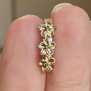 May include: A gold-coloured earring with three flower-shaped charms. The earring is held between fingers, highlighting the delicate floral design. The flowers are evenly spaced and have detailed petals.