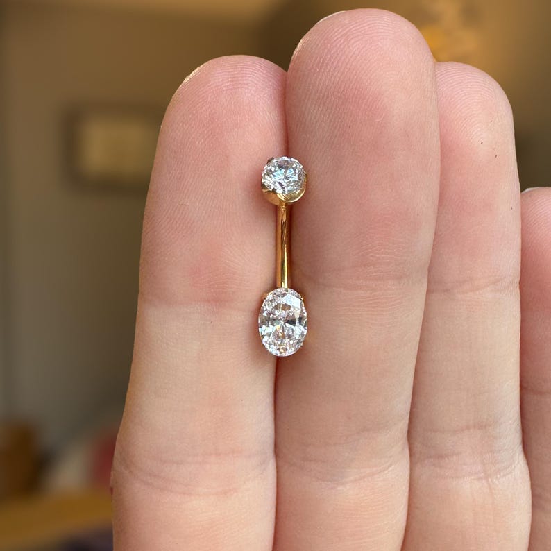 May include: A gold curved barbell body jewelry piece featuring a small round clear gemstone at the top and a larger oval-shaped clear gemstone at the bottom. The metal has a polished yellow gold finish, and the stones are set in secure metal prongs.