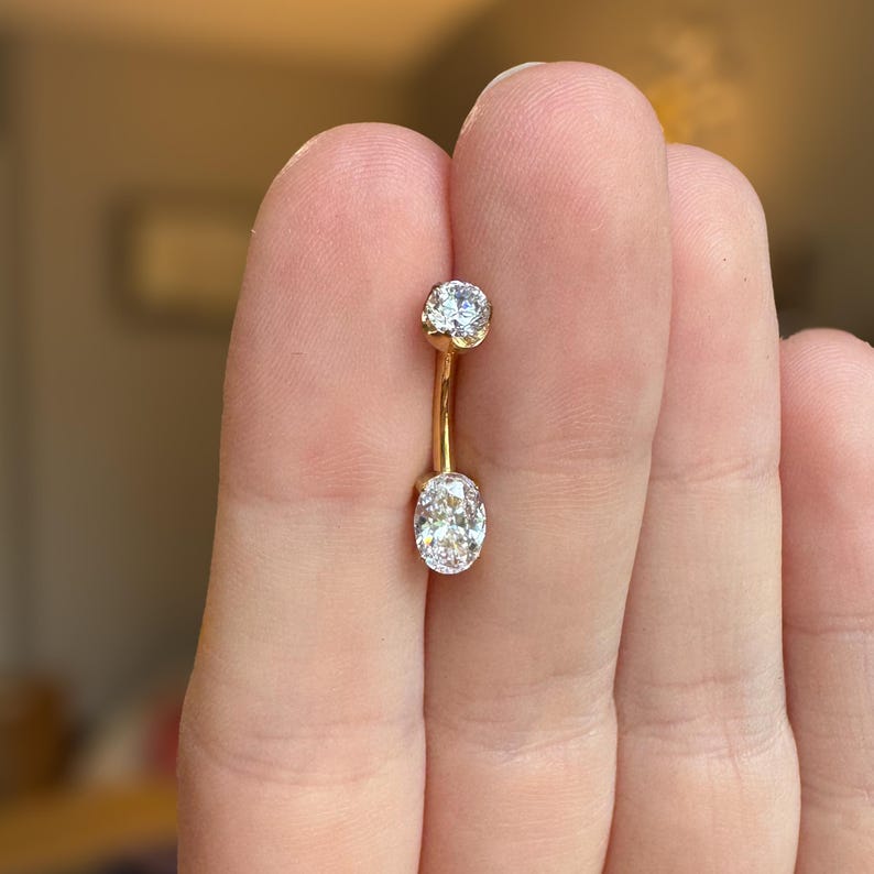 May include: A gold curved barbell belly button ring featuring a small round clear gemstone at the top and a larger oval-shaped clear gemstone at the bottom. The metal is polished gold, and the stones are faceted to reflect light clearly.