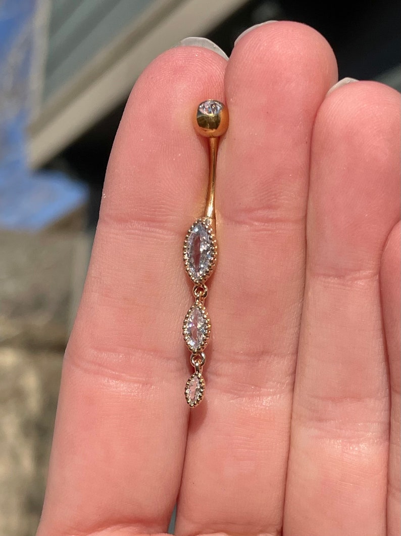 Puede incluir: Anillo de ombligo dorado con tres piedras claras en forma de marquesa.