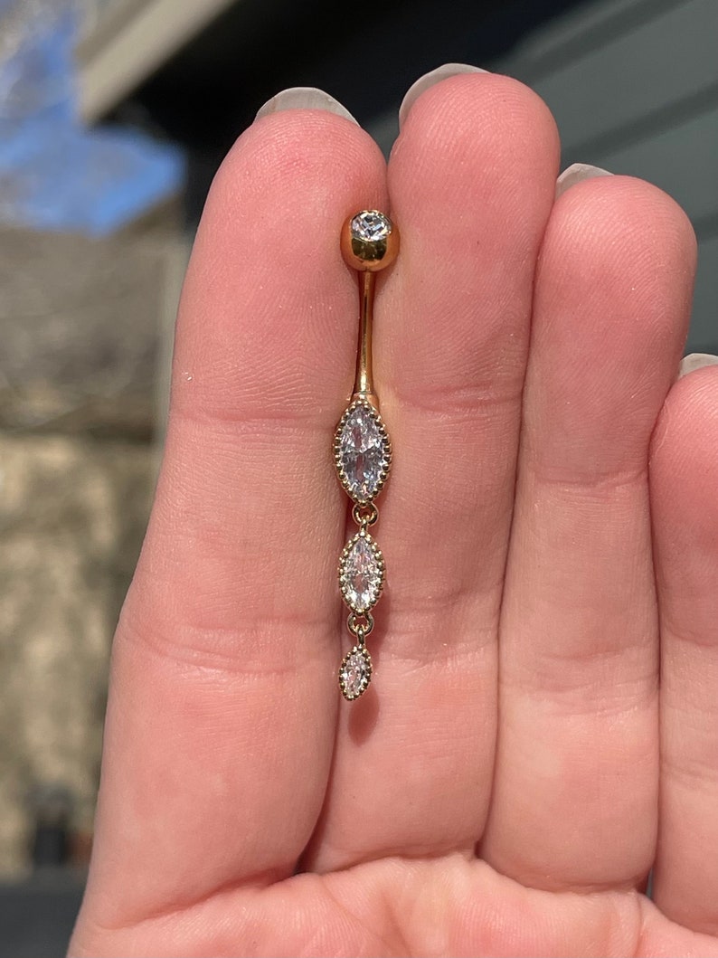 Puede incluir: Anillo de ombligo dorado con una cadena colgante de tres piedras transparentes en forma de marquesa.