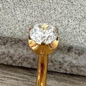 May include: Close-up of a gold-colored nose stud with a large, clear, round-cut diamond. The diamond is set in a gold bezel setting. The nose stud is on a gray, textured surface.